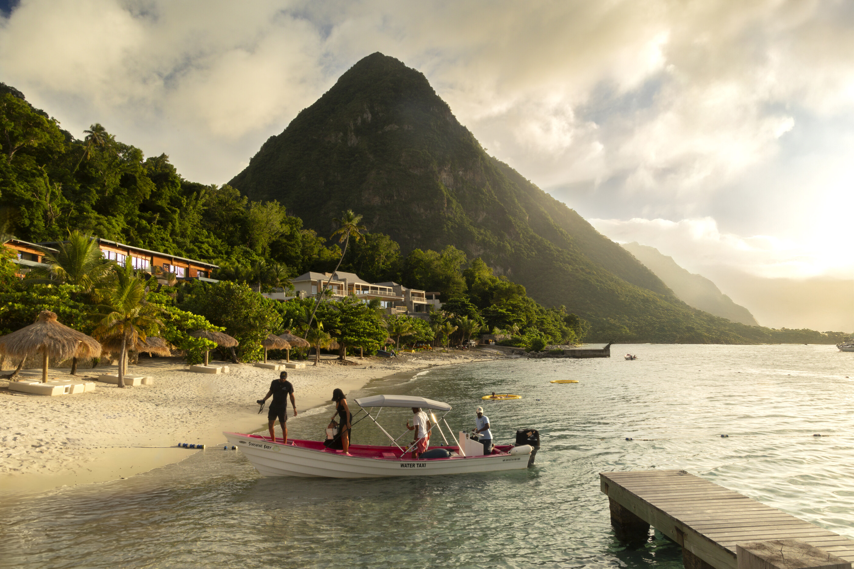 Two figures pulling a small wooden boat onto a beach in the Caribbean with a steep volcanic peak rising behind.