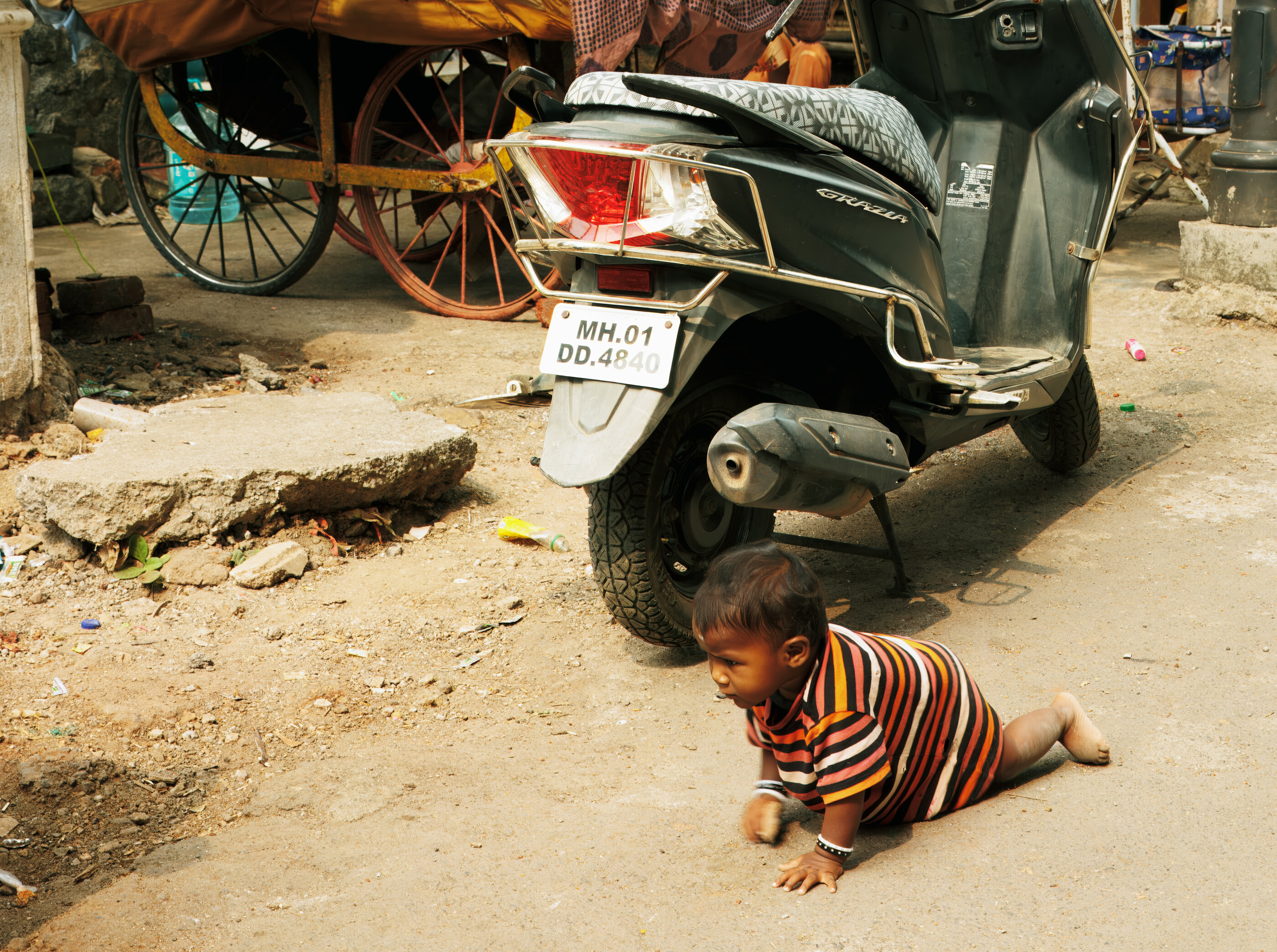 An Indian street with a parked motorcycle, a small dog in costume on the ground, old wooden buildings behind. The scene is dense and colourful, daily life in India. Travel-as-subject.