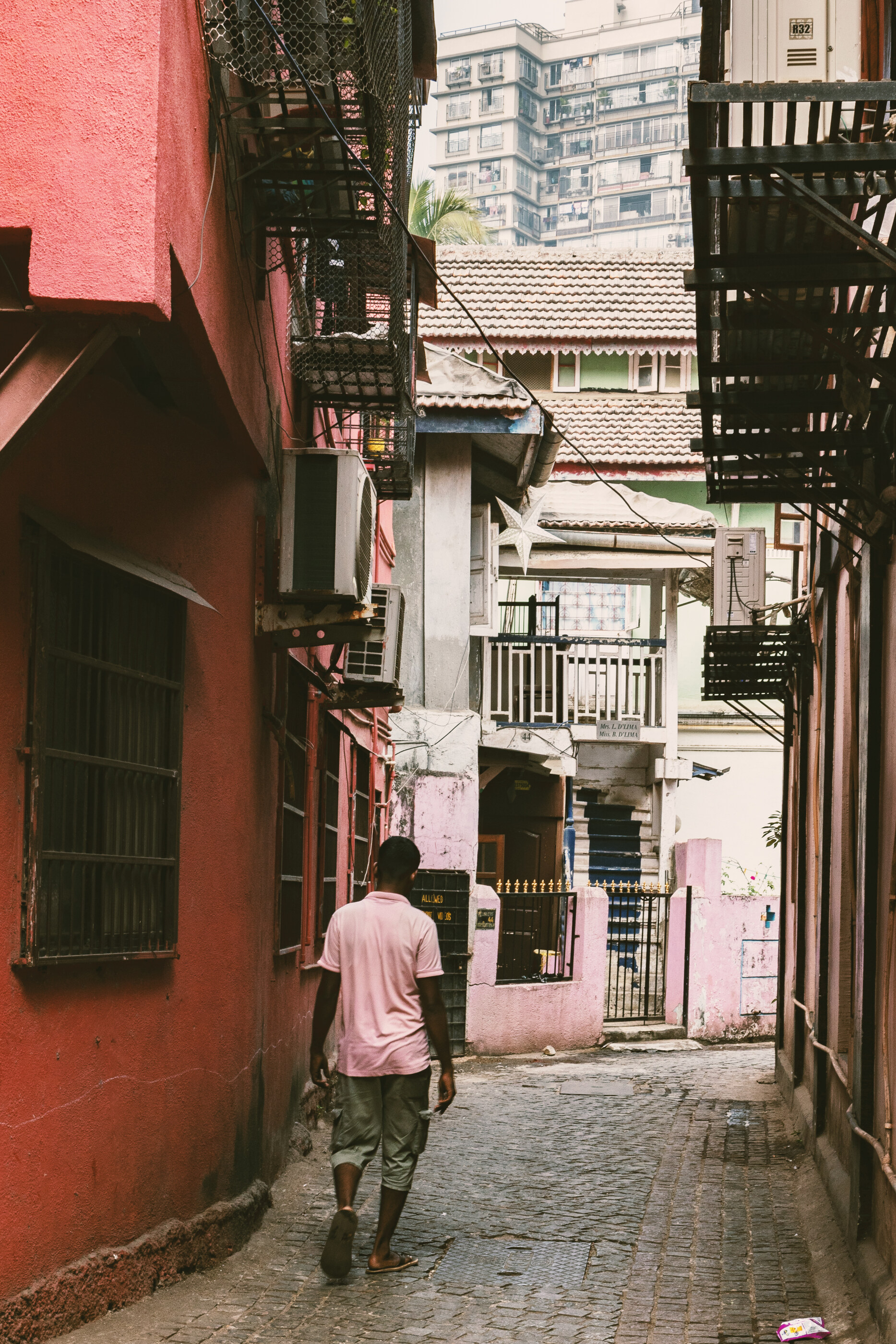 A figure walking down a sun-soaked Indian alley between pink and red buildings, photographed from behind.