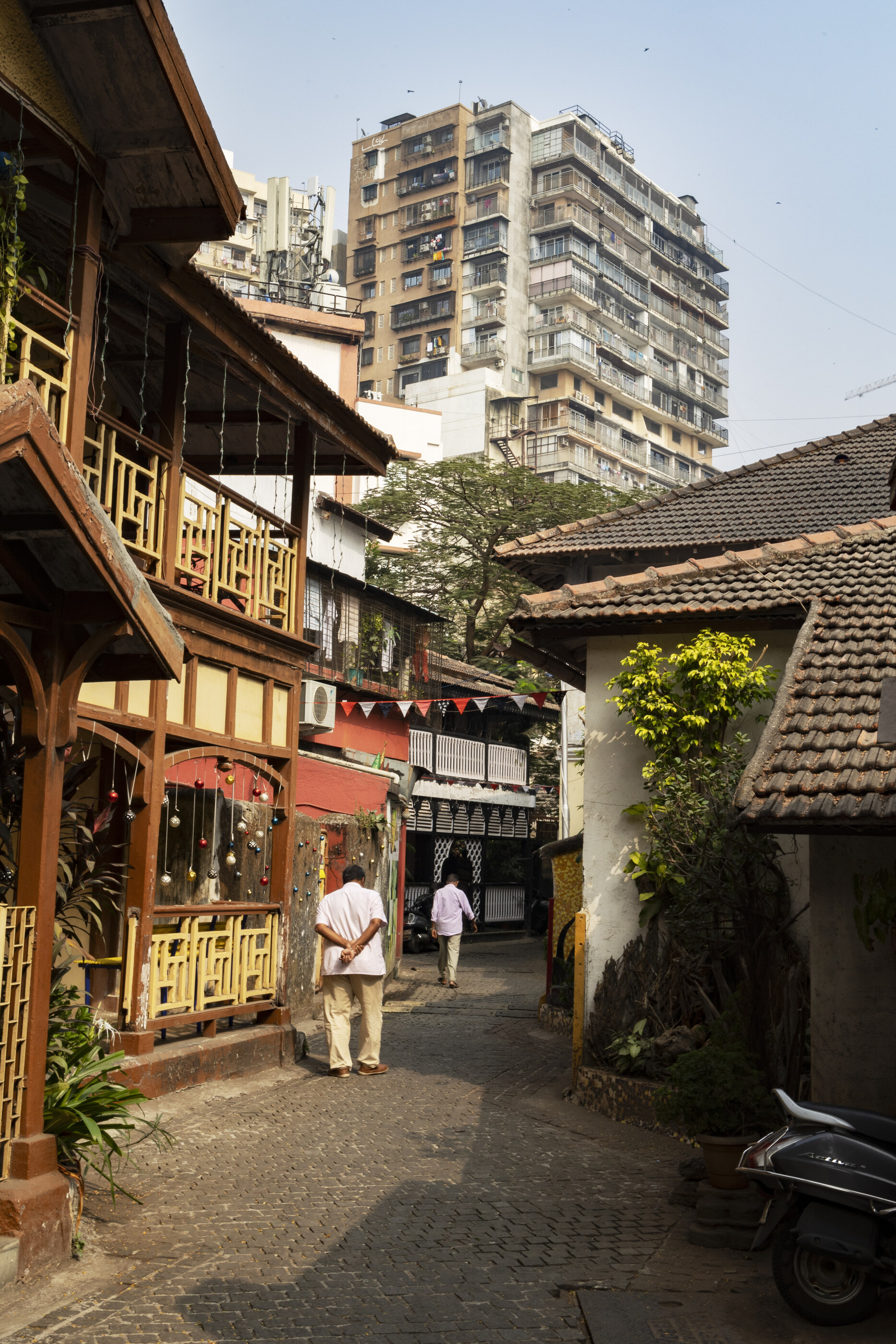 An Indian alleyway with cracked walls, a few small figures and shopfronts under a strong shaft of sunlight. Densely textured; daily Indian street life is the subject.