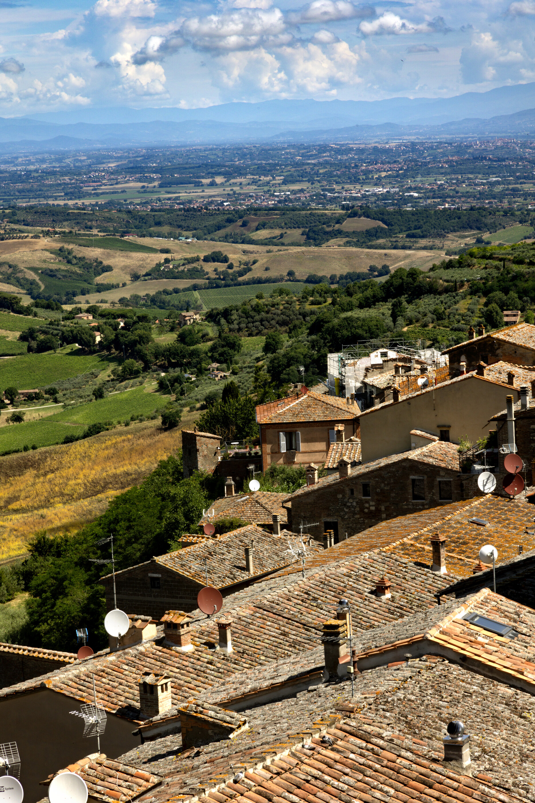 A wide Italian valley landscape with a hilltop town clustered to the right and rolling fields beyond. The land dominates; the town is integrated into it. Travel-as-subject: Italy.