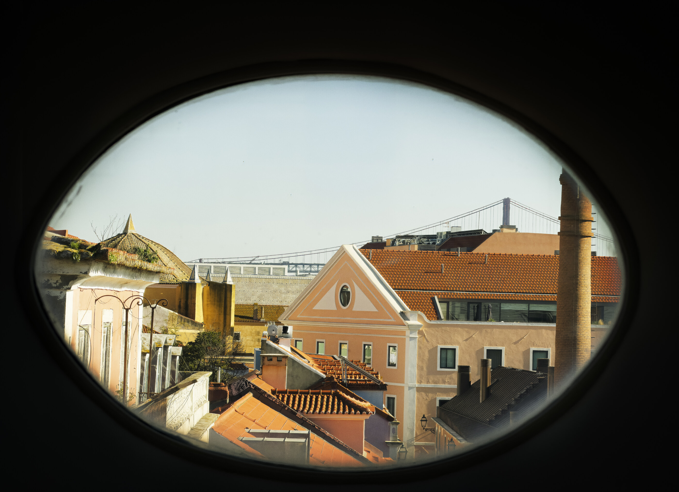 A view through a circular porthole window onto orange rooftops and a city beyond. No people. The frame-within-a-frame and the built city seen through it is the subject.