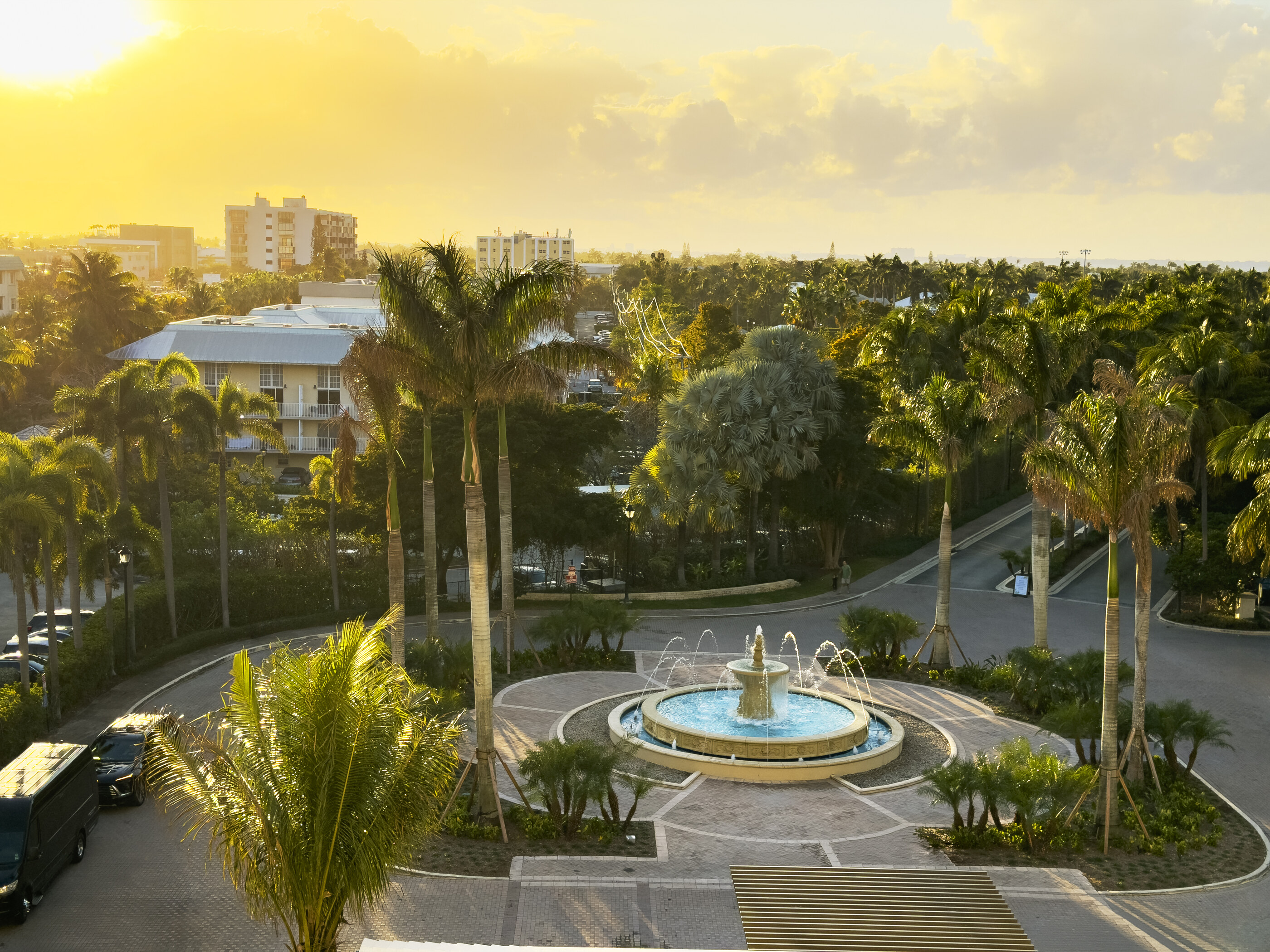 A tropical resort courtyard with tall palm trees, a fountain, and a low building wing in golden light. No dominant person. The composed architecture and landscape of the resort is the subject.