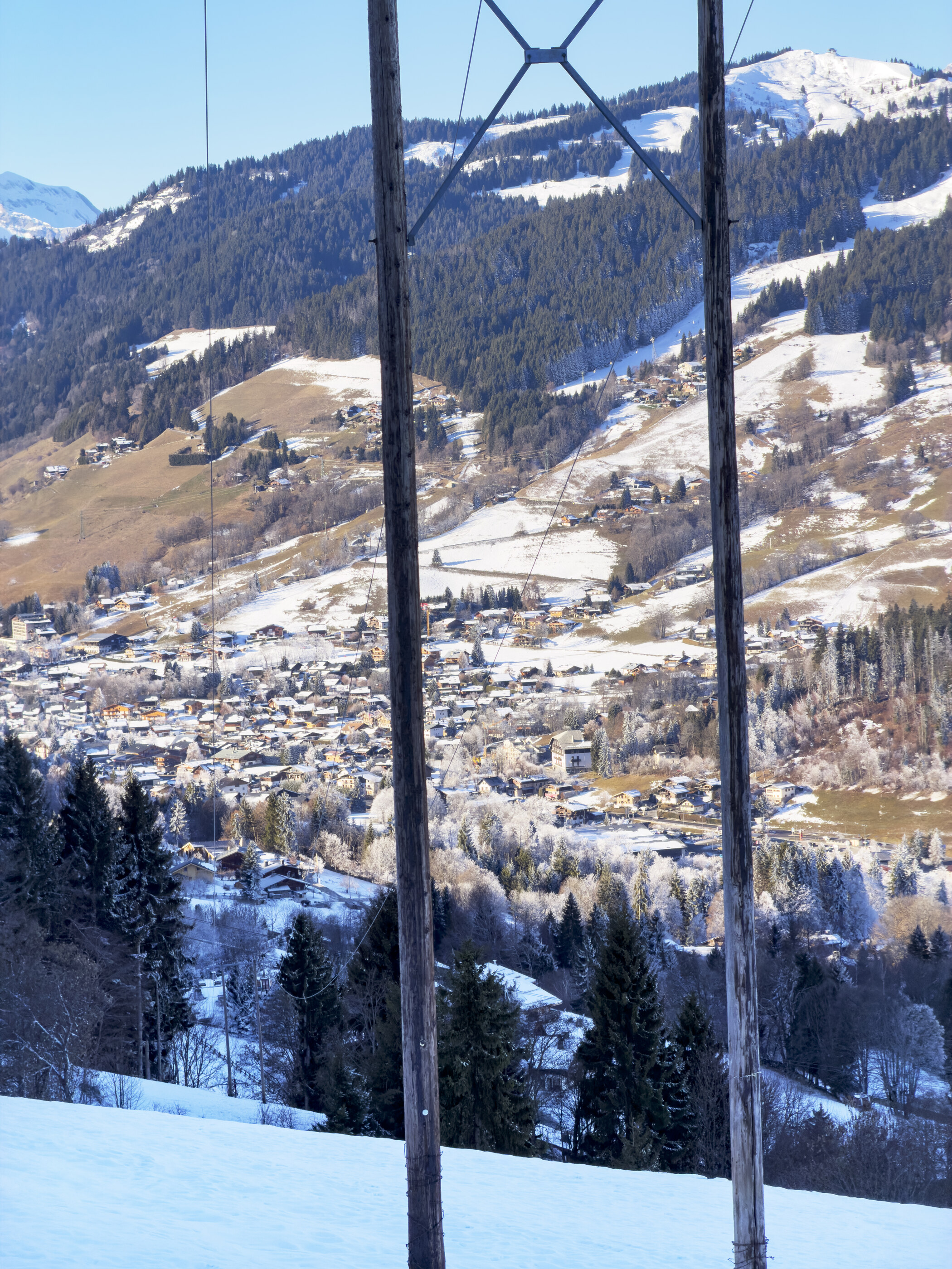 An alpine village seen in winter, blanketed in snow, with electrical lines and a partial mountain framing the scene.