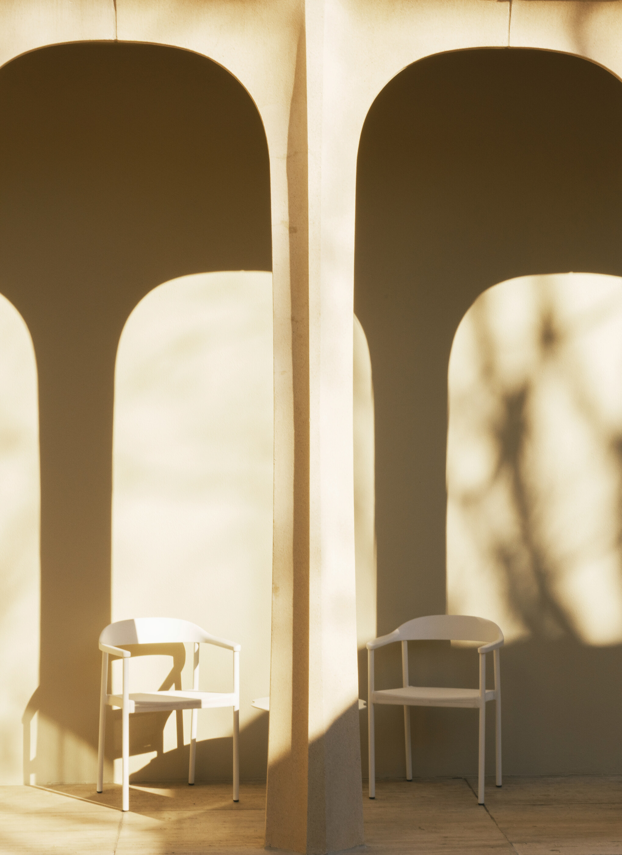 A sunlit interior with two arched openings, two slim chairs, and tree shadows on a cream wall. No people. The interior architecture — arches, light, chairs — is the subject.