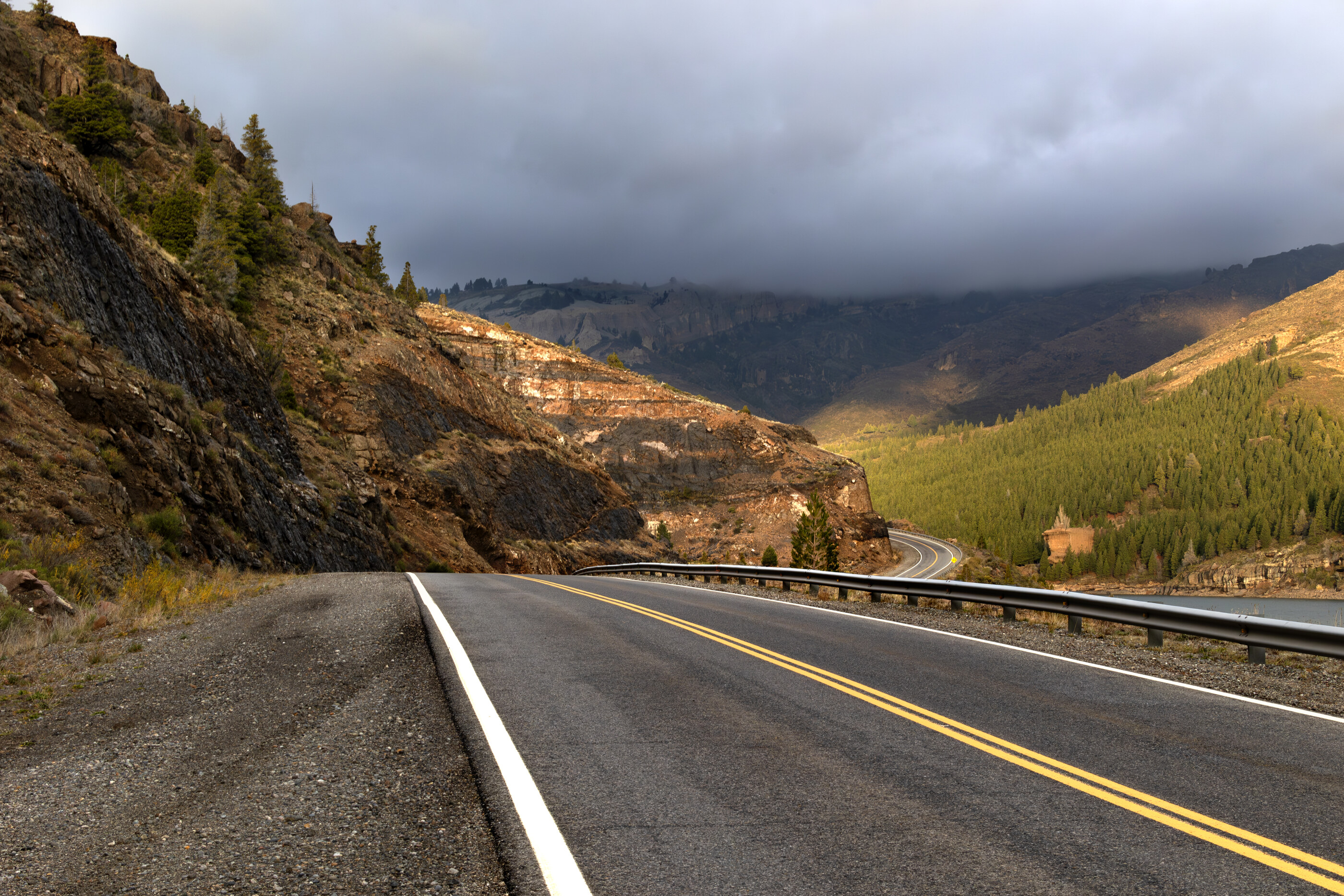 A winding mountain road descends into a hazy valley under storm clouds, no vehicles visible. No people. Vast quiet road in landscape.