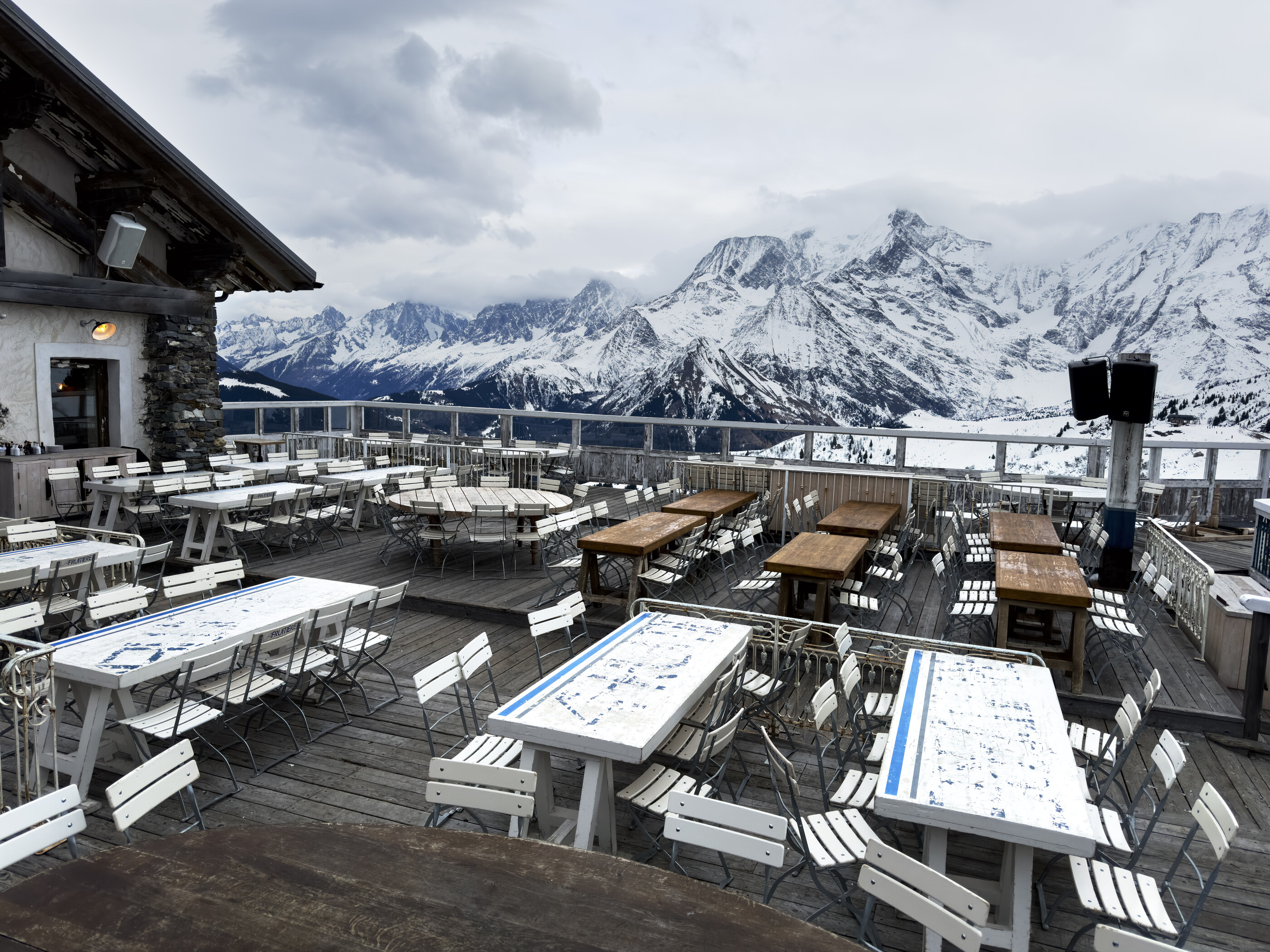 A mountain restaurant's terrace with empty wooden tables under a snow-capped peak backdrop, late winter daylight.