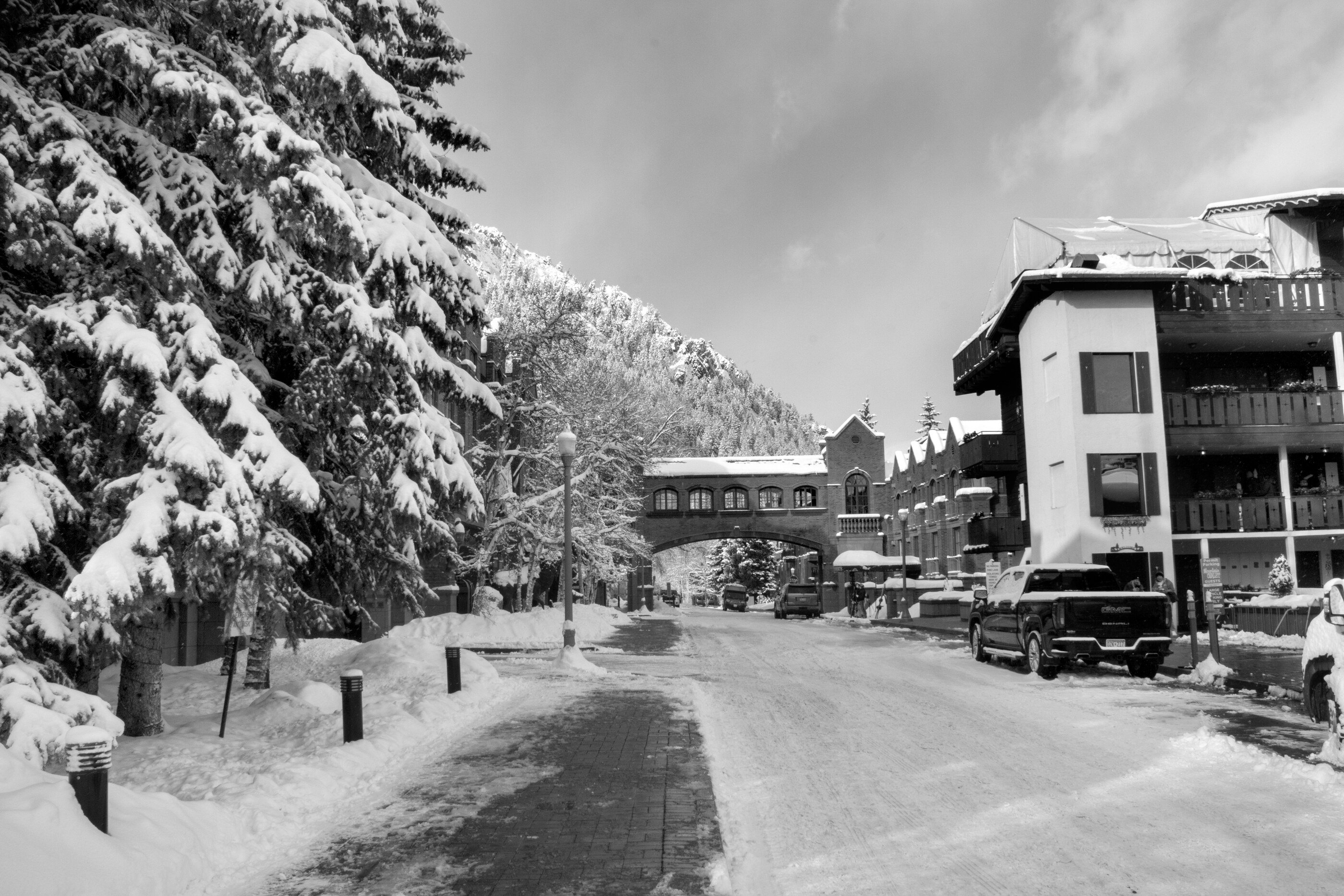 A snow-covered road in black-and-white with cars and houses in soft winter light, no visible person. Quiet snowy street; slow time.