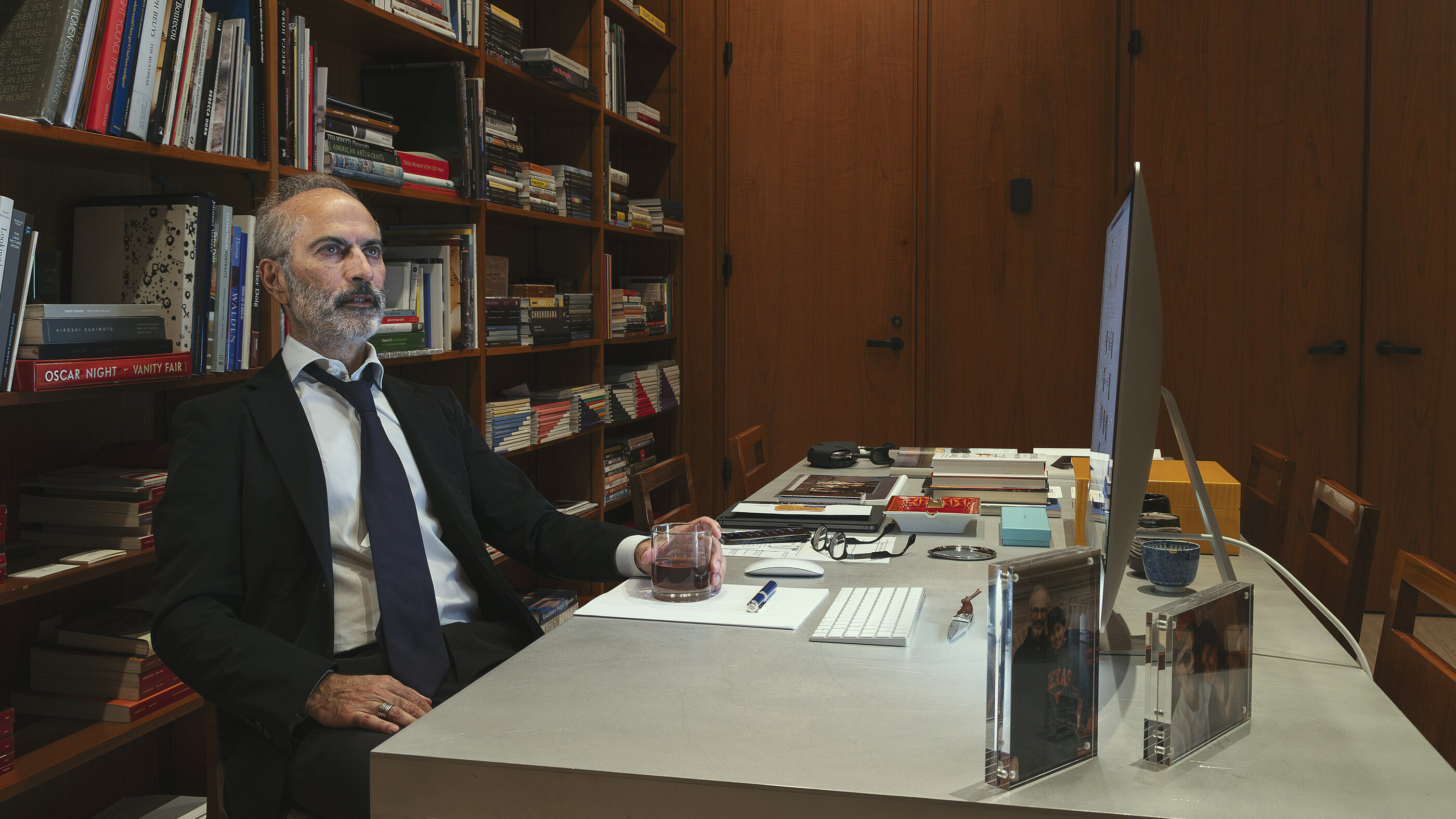 An older man in dark suit sits at a desk in front of a wall of red-bound books, mid-30s environmental portrait. He is the subject; the books are context. Posed and lit.