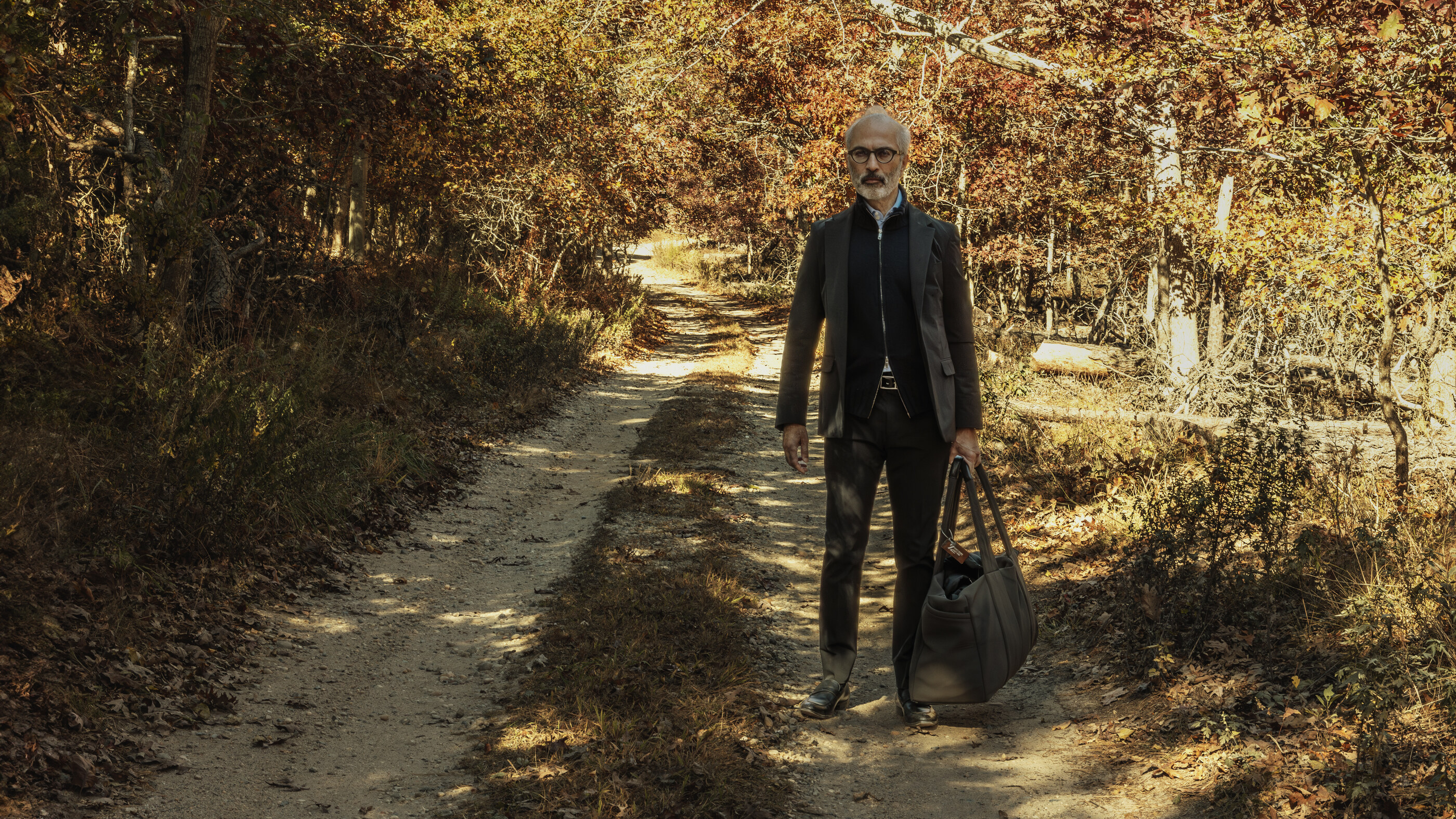 A single figure with their back to the camera, walking through a sunlit urban courtyard or alley with bag in hand.