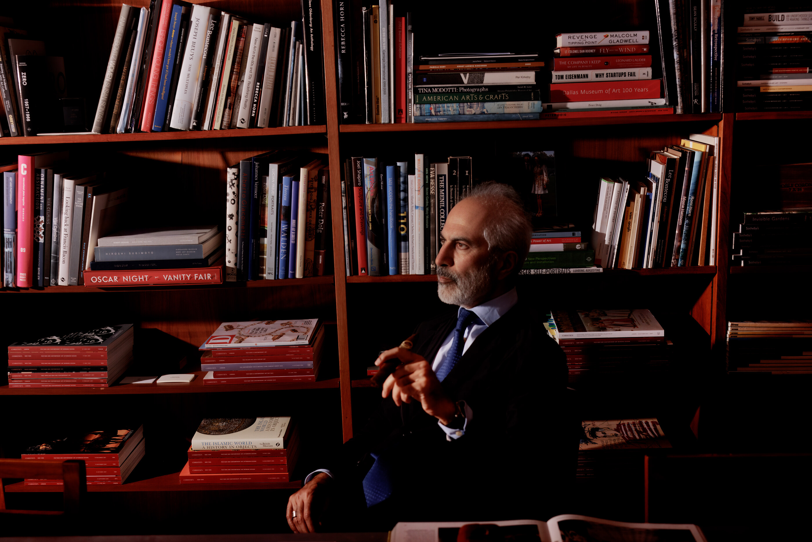 An older man in a dark suit sits in a leather armchair in front of a red bookshelf, posed environmental portrait.