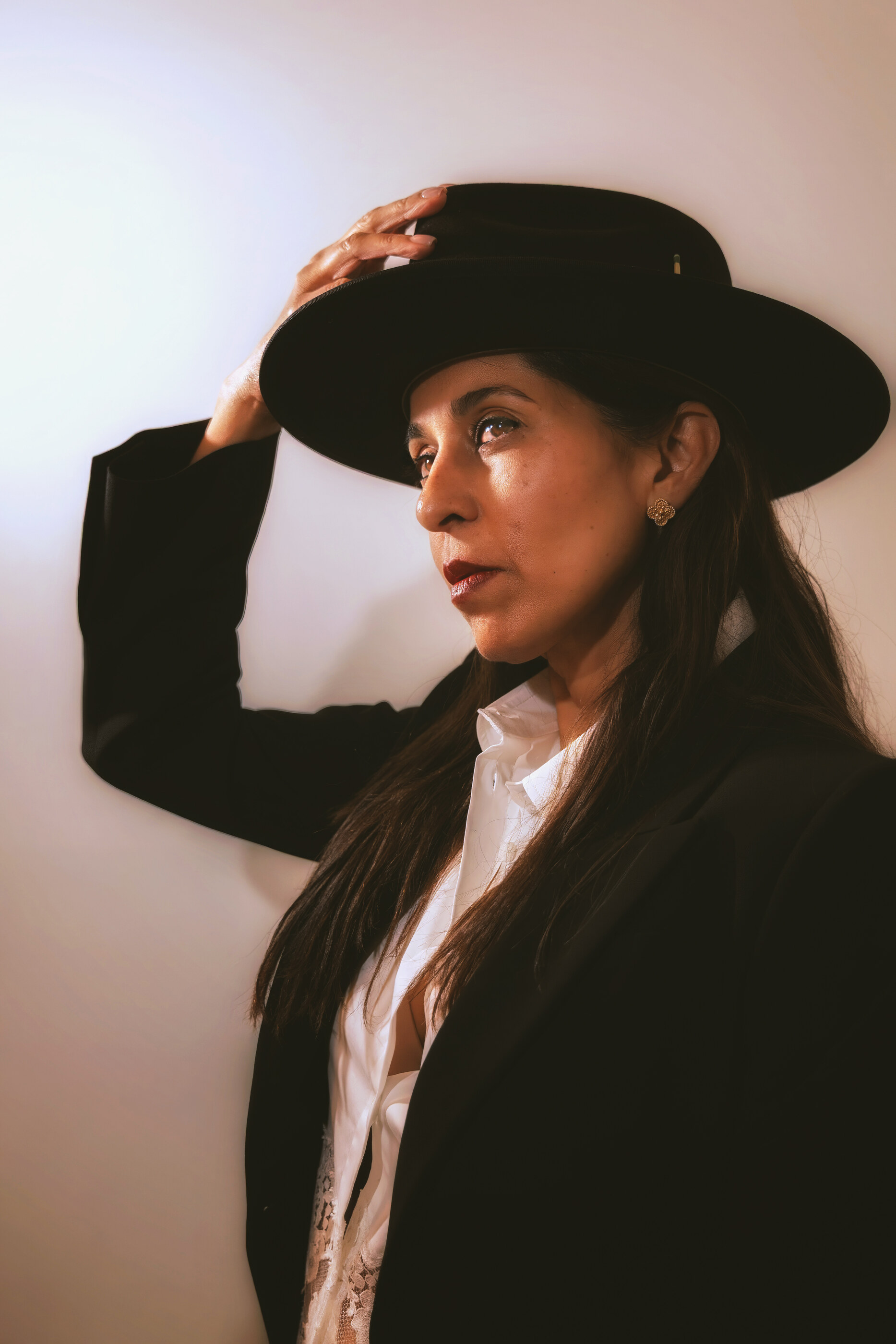 A young woman in a black coat and wide black hat, three-quarter studio-style portrait, looking up to the side. She fills the frame and is the unambiguous subject. The lighting is soft and warm.