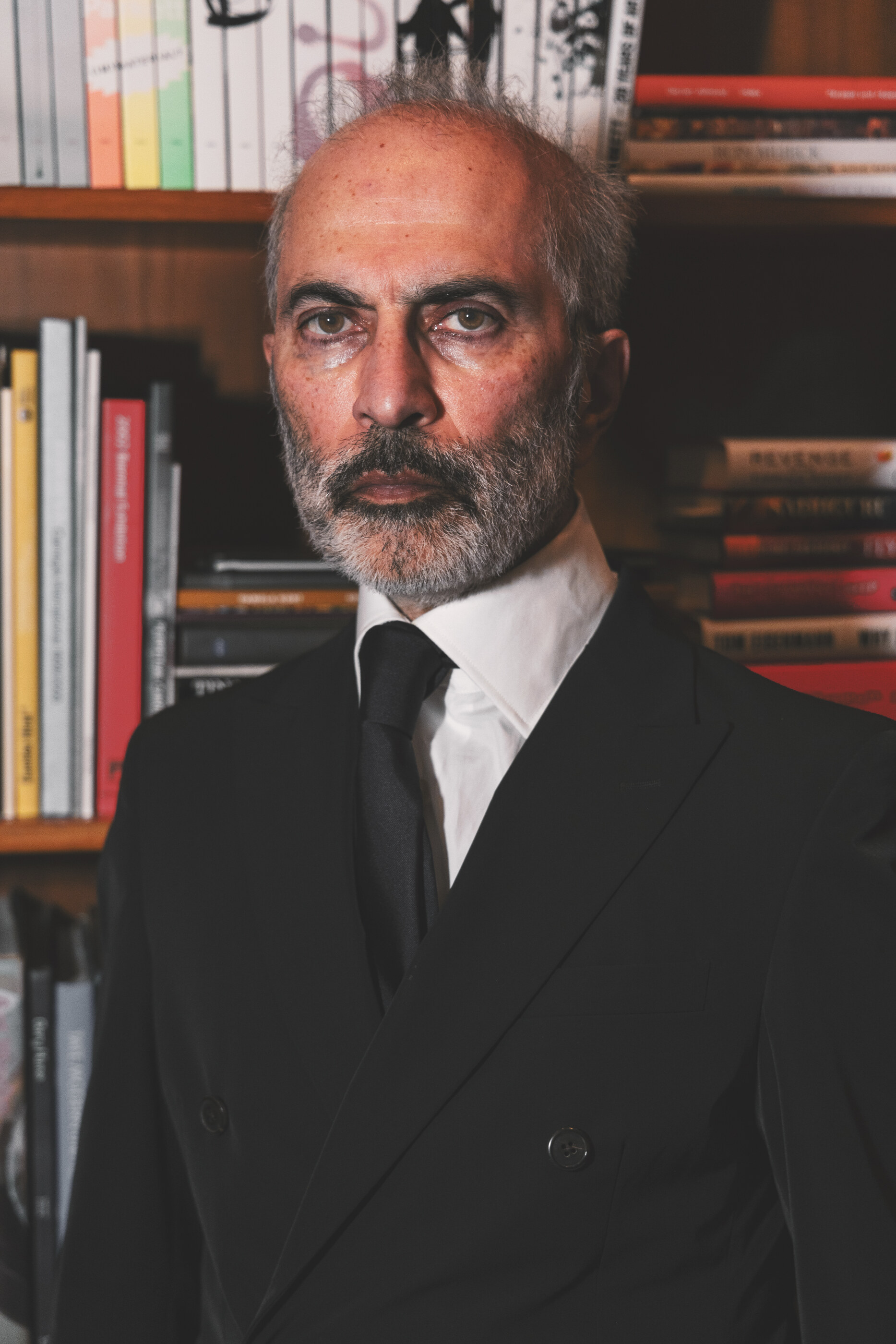 A close half-length portrait of an older man in a black suit against a backdrop of books. He fills the frame; the books are blurred context. Classical posed portrait.