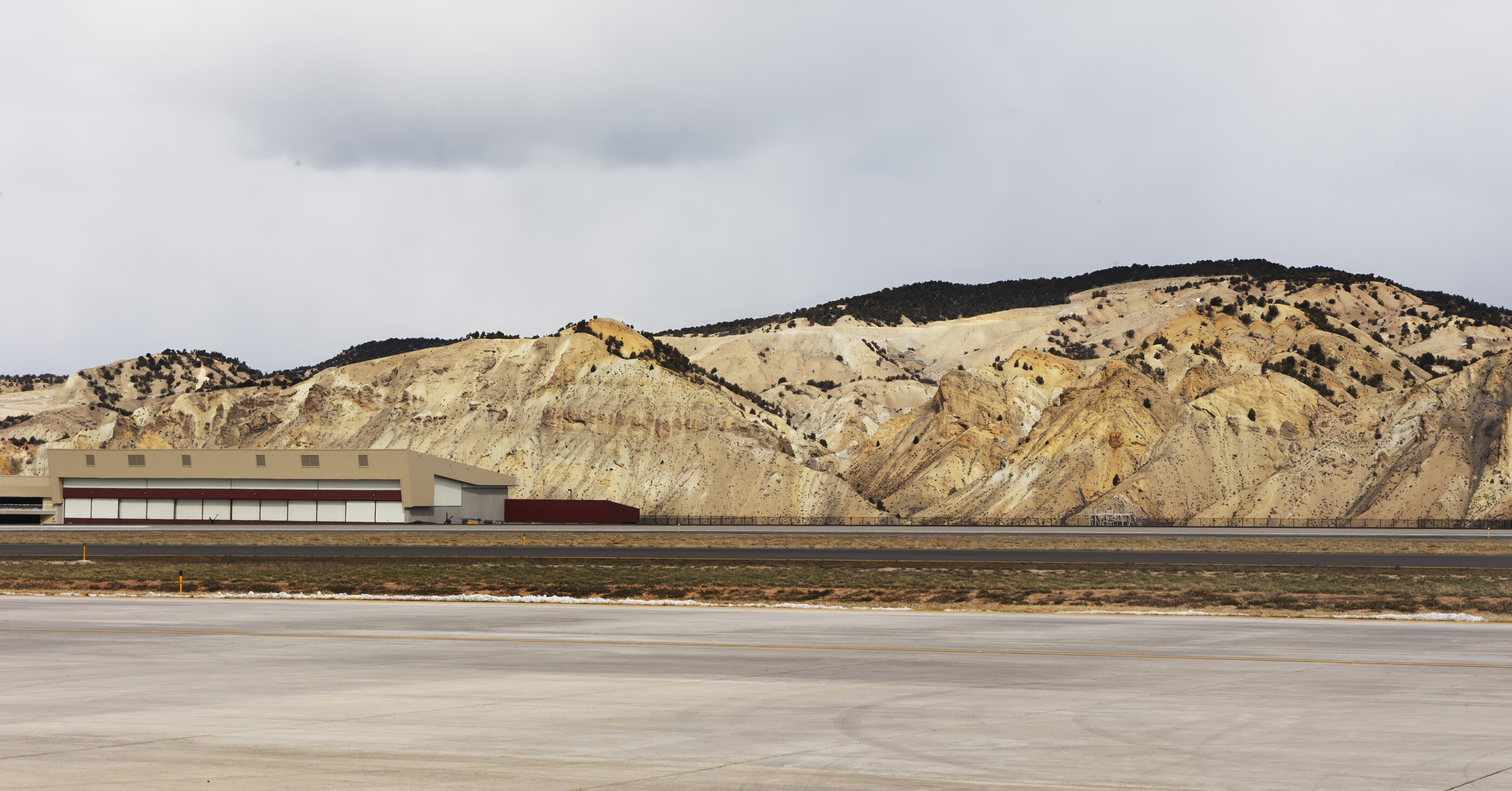 An airport runway in the foreground with a mountain range beyond and storm clouds overhead. No prominent person. The wide vista combining built and natural land is the subject.
