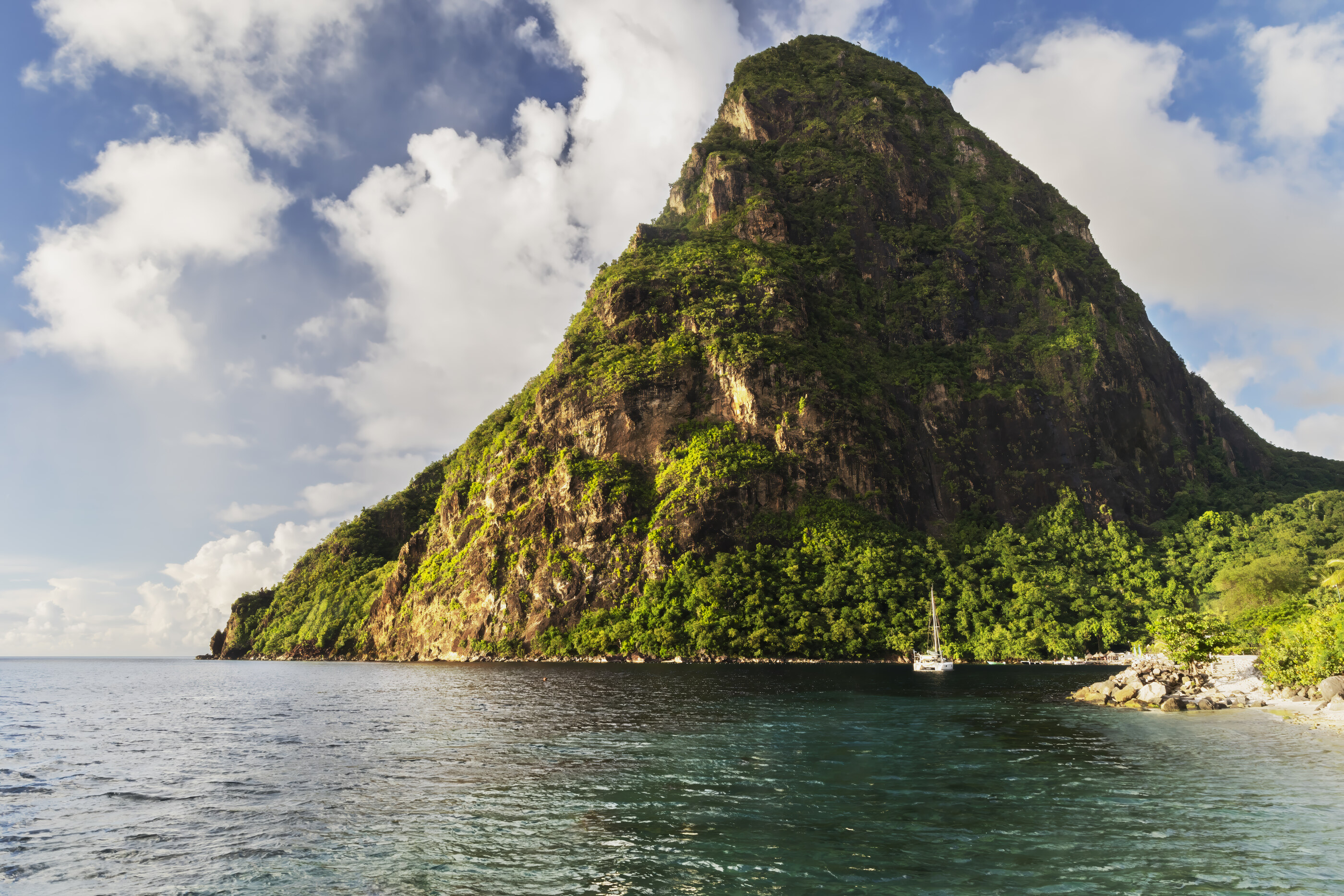 A Caribbean coastal scene with a steep green island peak rising over turquoise water and a small boat. No dominant person. Landscape at scale.