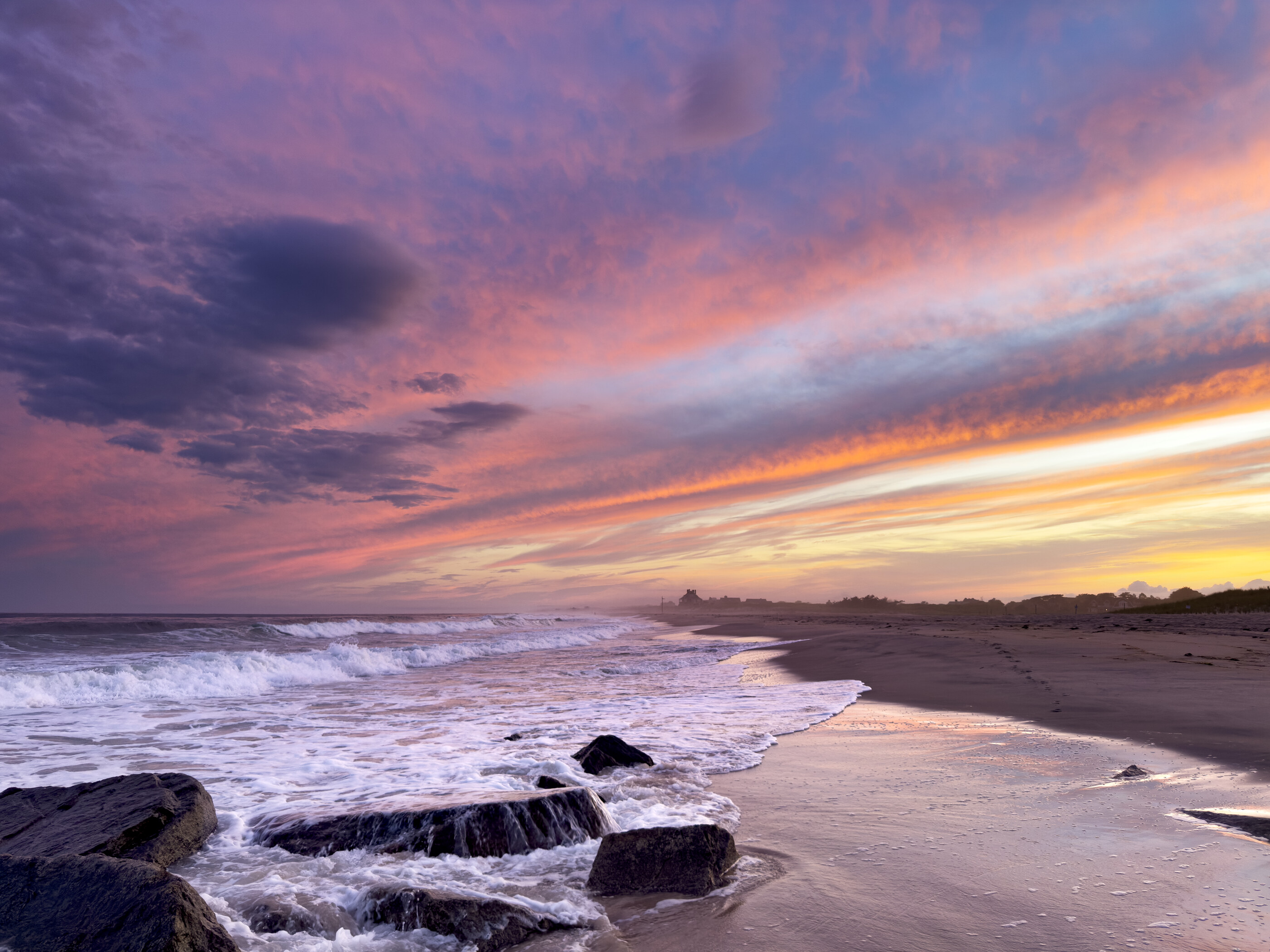 A sunset over a beach with breaking waves, dramatic clouds streaked across the sky. No people in frame. The light over the coast is the photograph; place at scale.