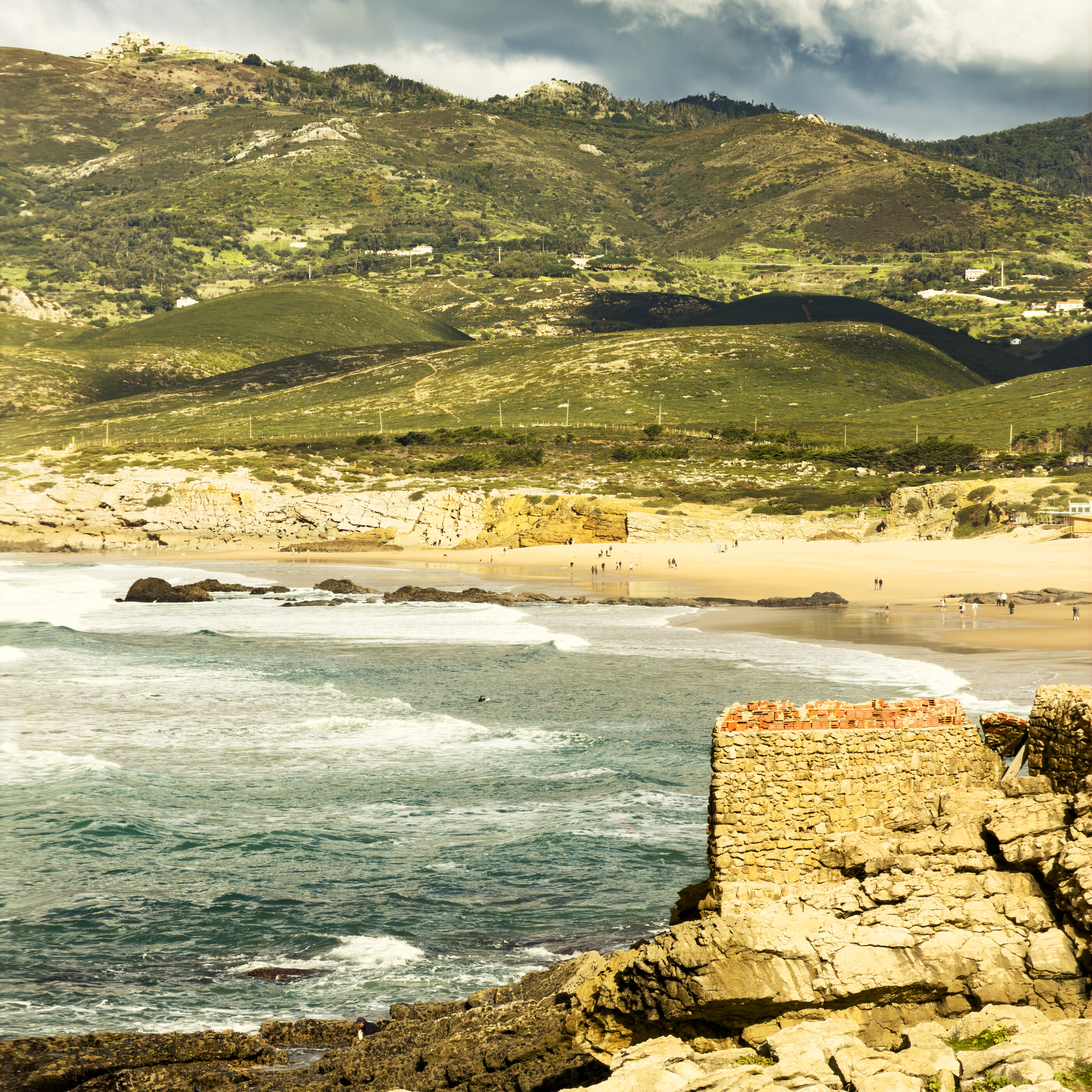 A wide Atlantic coastline with cliffs, breaking surf, and rocks in foreground (Cascais, Portugal). No dominant person. Landscape at coastal scale.