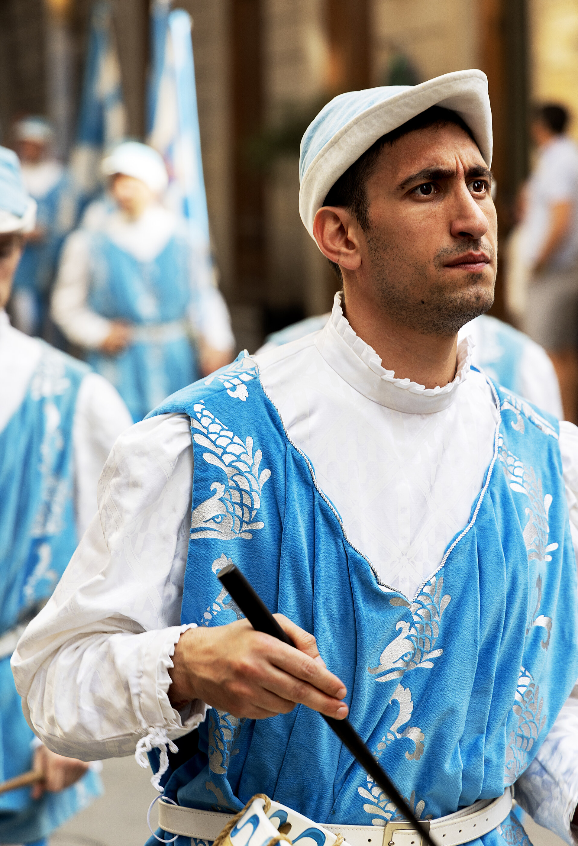 A man in a blue-and-white religious or ceremonial costume holding a drum stick or brush, mid-procession in a crowd.