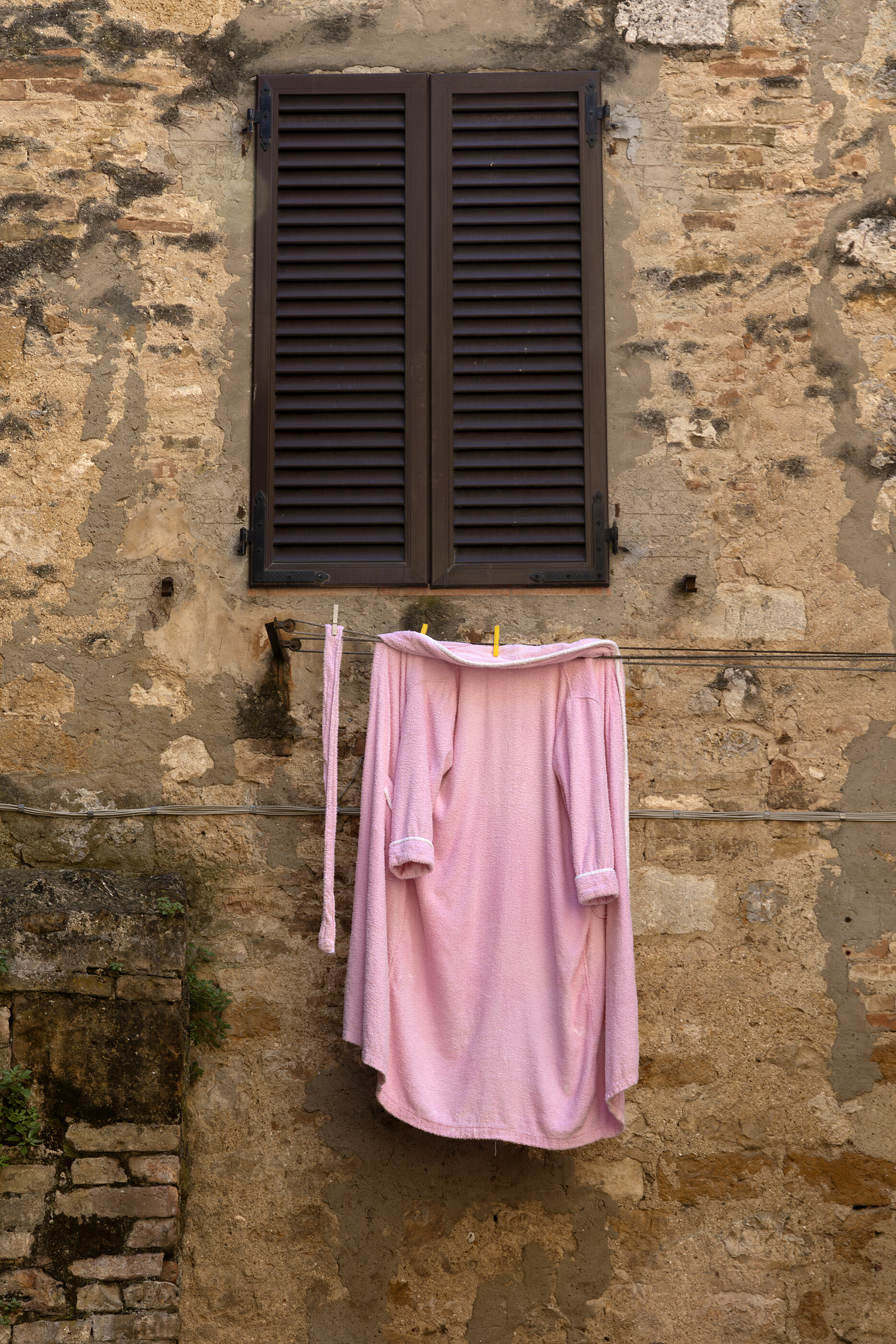 A pale pink robe / cloth hangs from a stone wall under a closed wooden shutter, in golden side light. No people. The robe as a noticed object is the subject — a found composition.