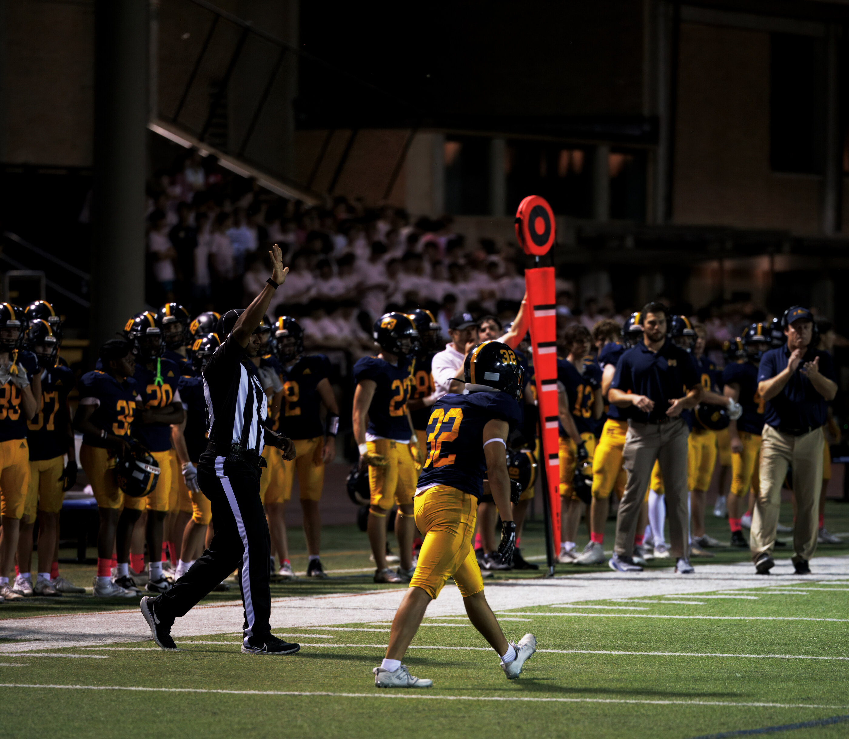 A football team and officials gather near the end zone or sideline, multiple players in pads. Sports context.