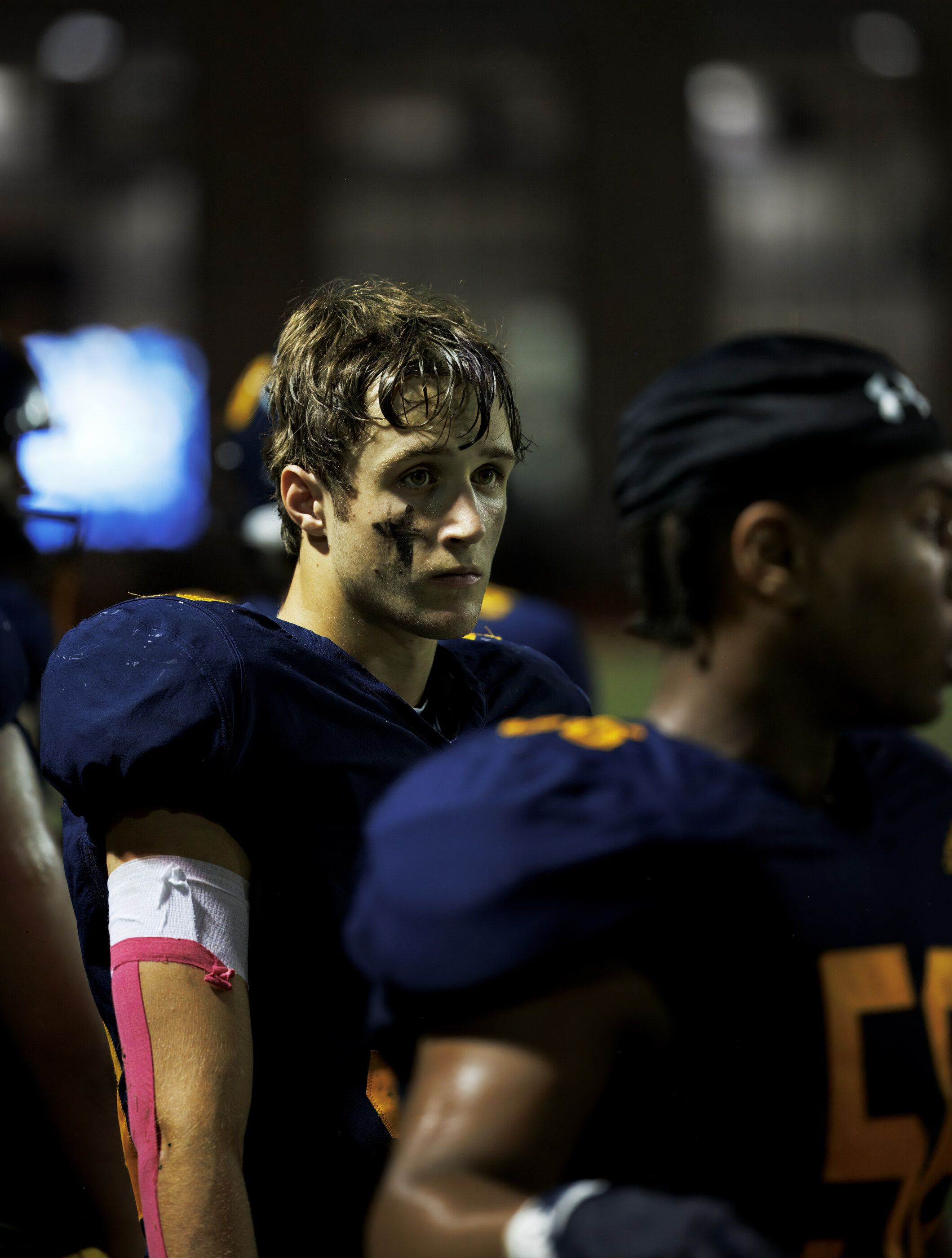A close shoulder shot of two football players on the sideline, helmets visible, in low evening light. The athletes and game uniforms identify it as American football — sport in context.