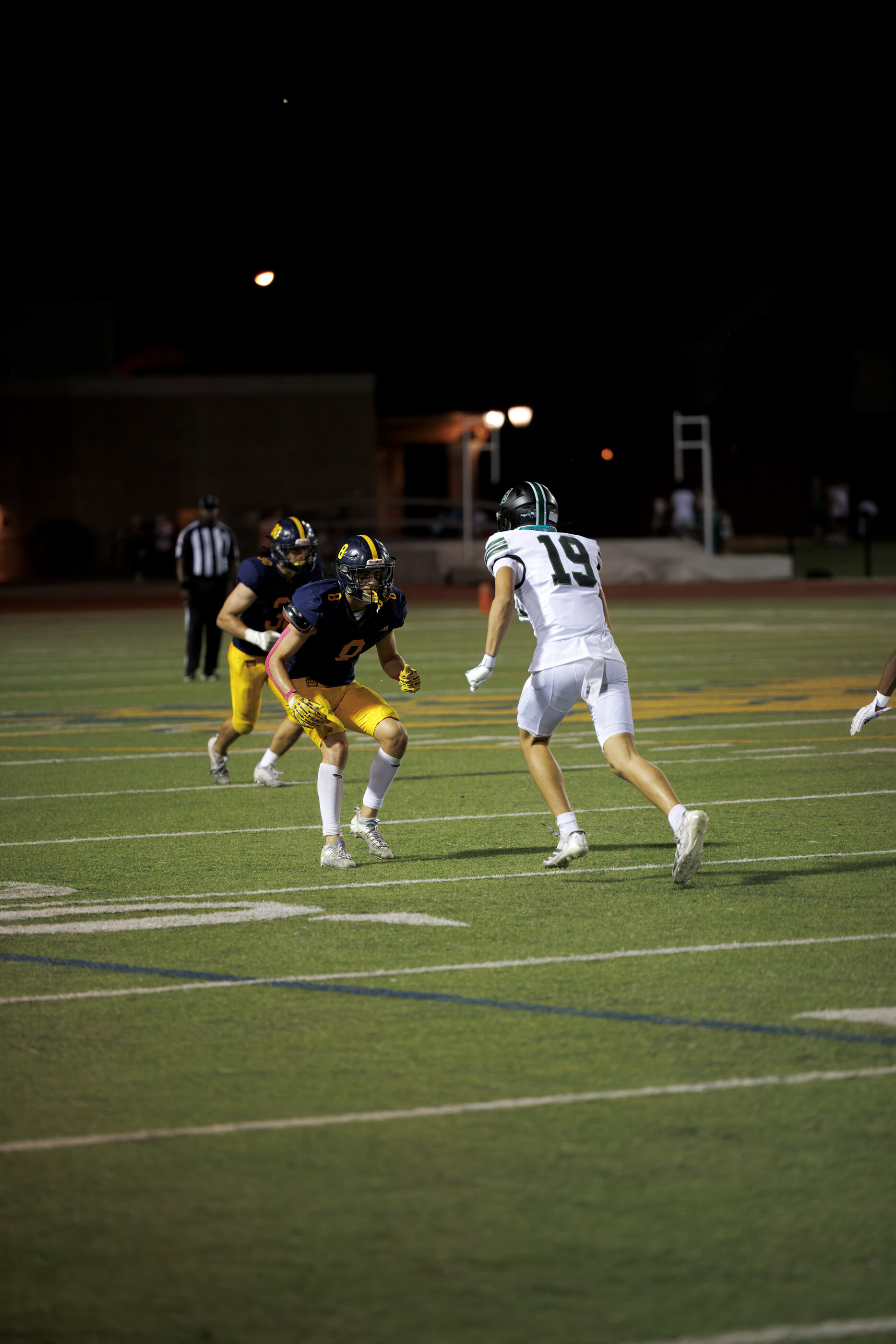 Football players running on a field under stadium lights at night, multiple figures in motion. Athletic activity is the subject.