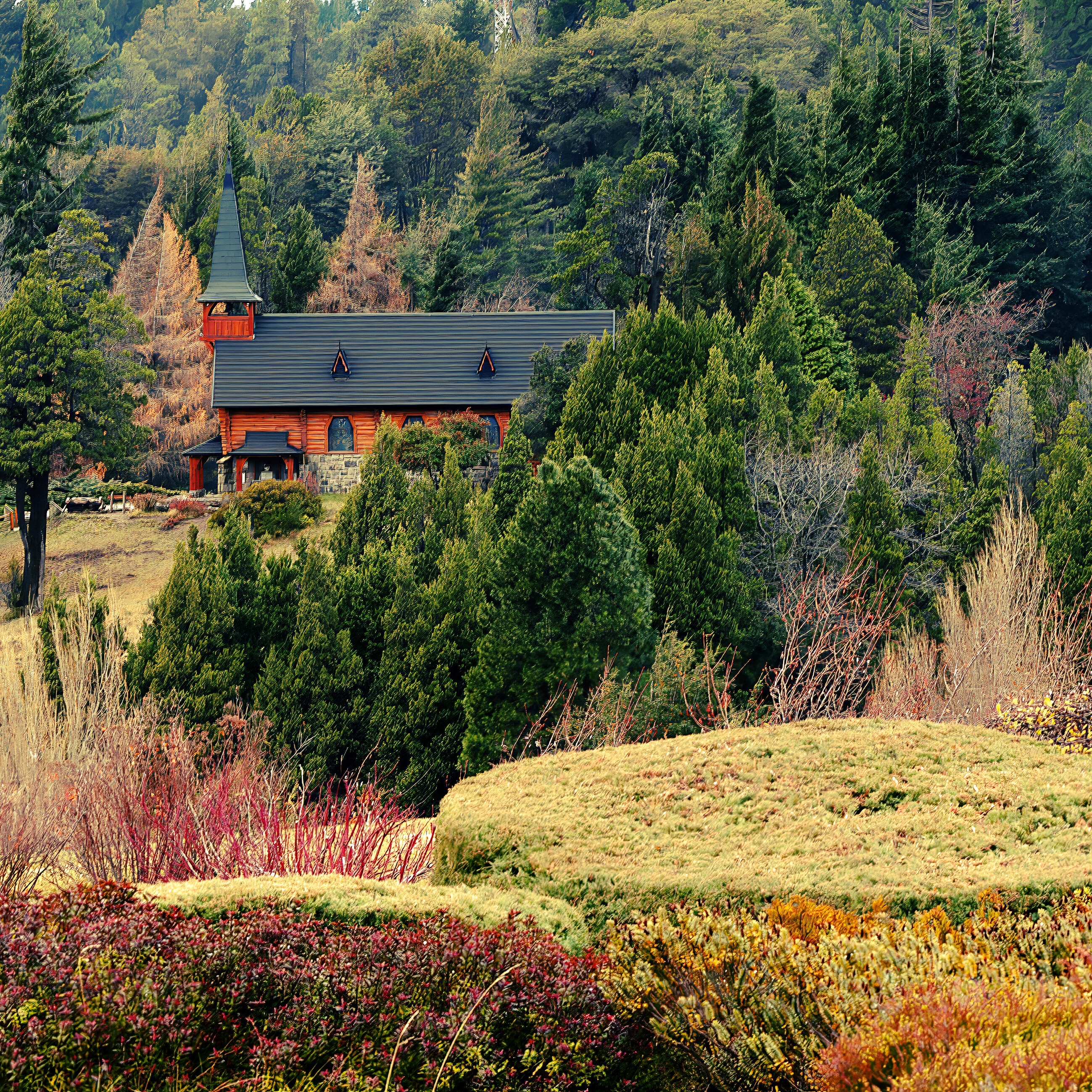A small red house in an autumnal meadow with mountains and forest behind, soft overcast light. No people. The scene is quiet, slow, rural; the house and its setting are the subject.
