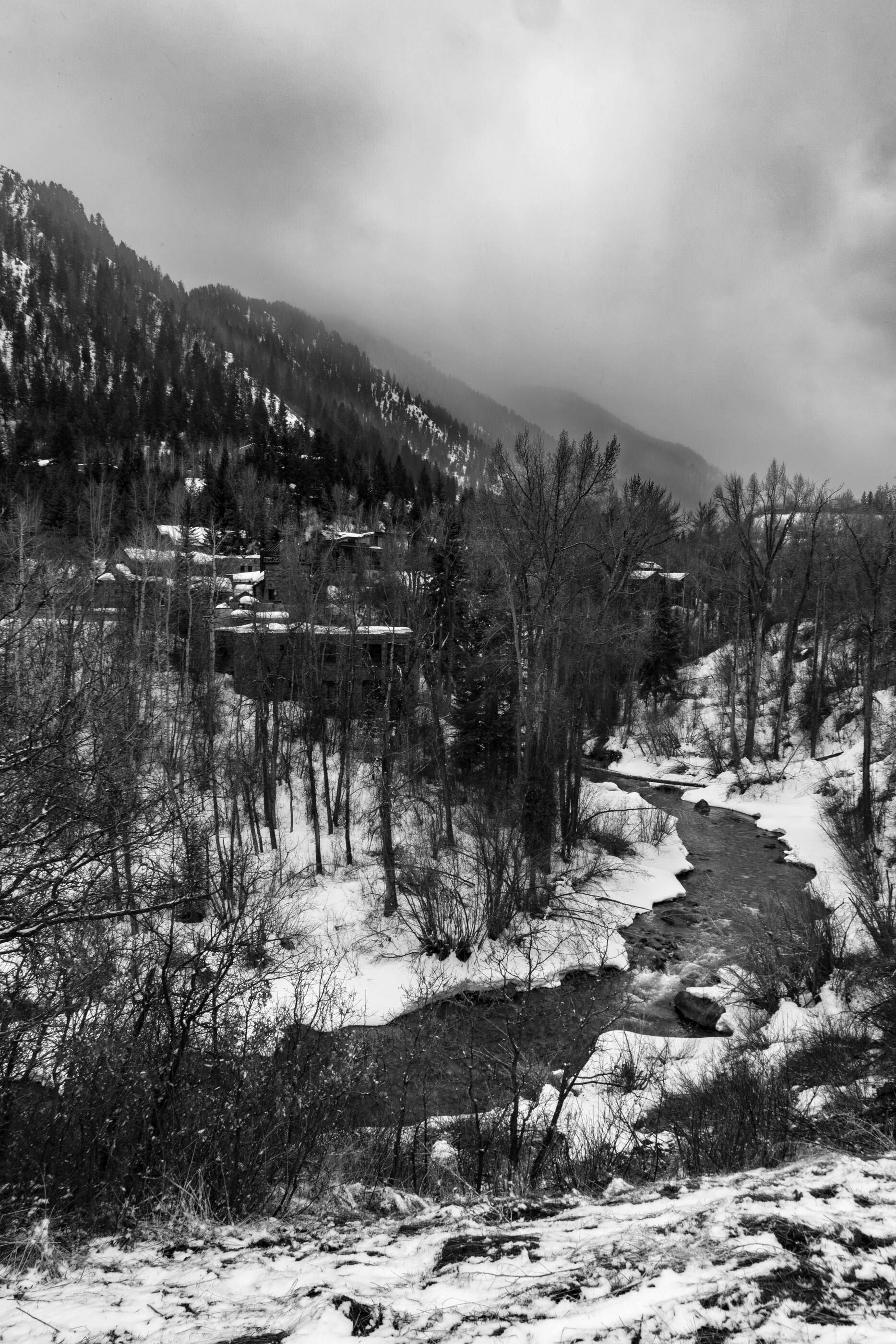 A winter stream in black-and-white, leafless trees and snow on the banks. No people. The image is quiet, contemplative, slow — a rural winter watercourse.