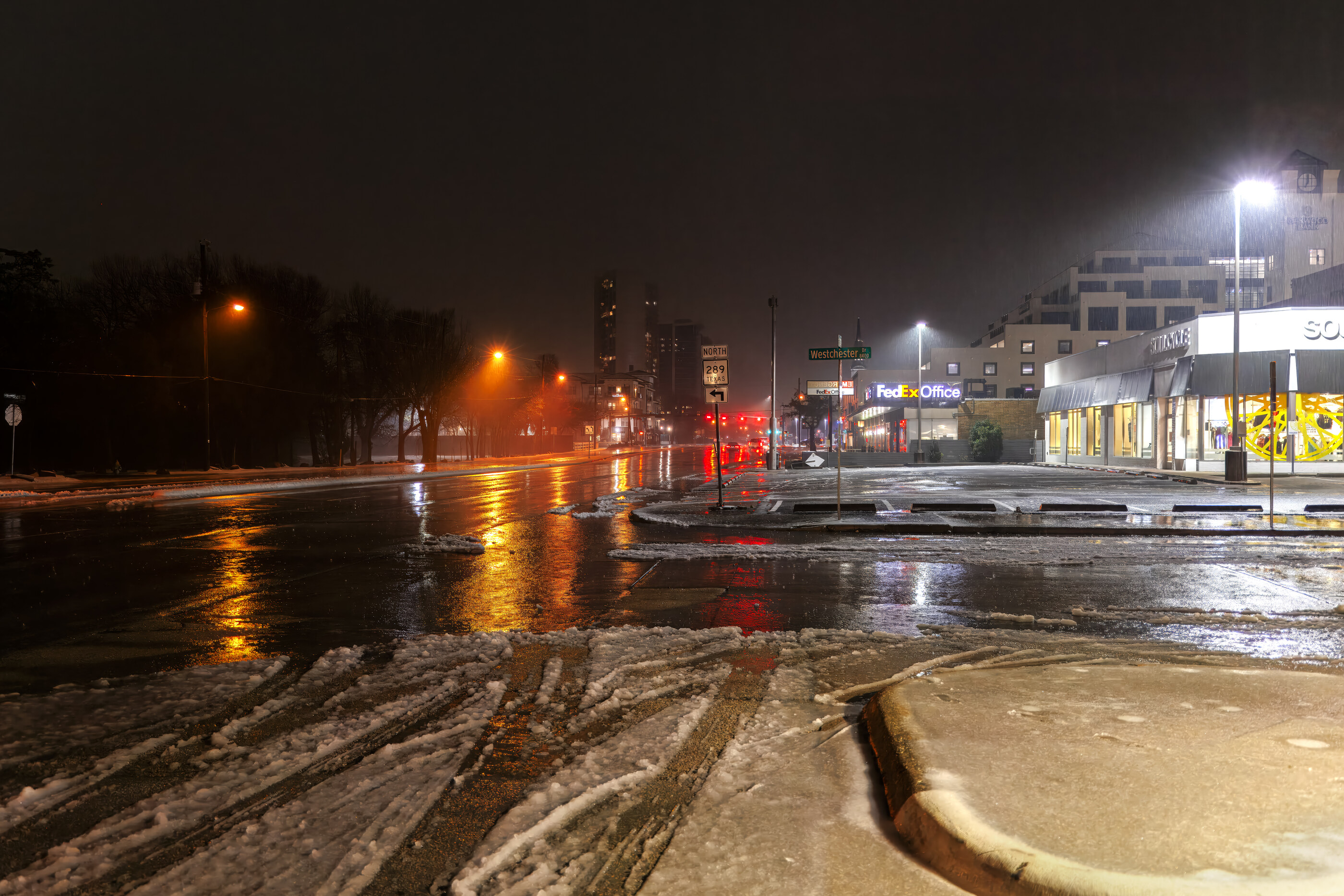 A wet road at night with car headlight reflections, urban edge in distance. No visible person. The photograph is about the wet, glowing night street — urban motion implied.