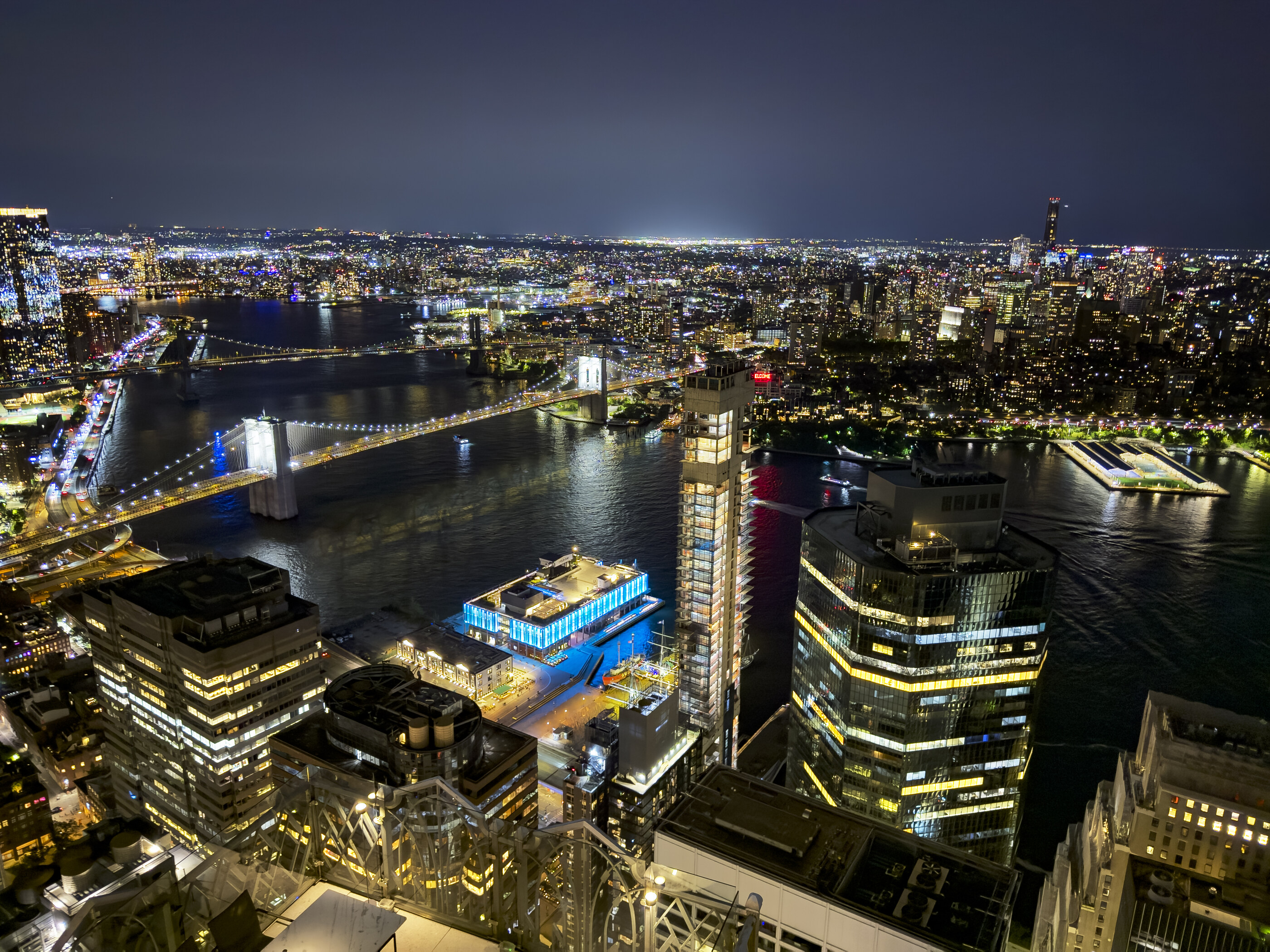 Another nighttime city skyline shot from above with water and a marina in the foreground, towers densely lit. No people. The urban scene at scale is the subject.