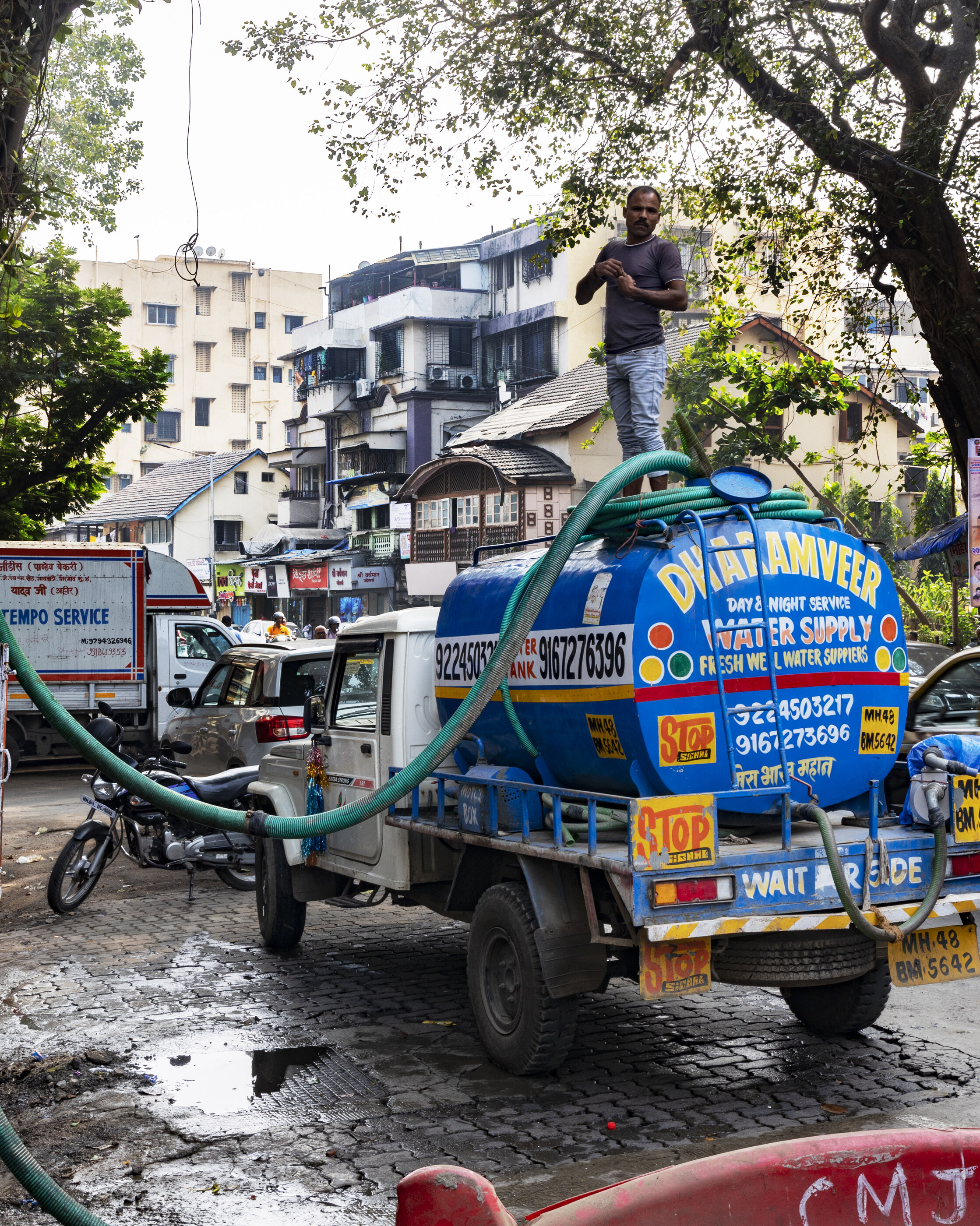 A blue Indian auto-rickshaw or van plastered with signs sits in the middle of a wet street, a figure standing on top doing some work. Plural activity; Indian transit and signage is the subject.