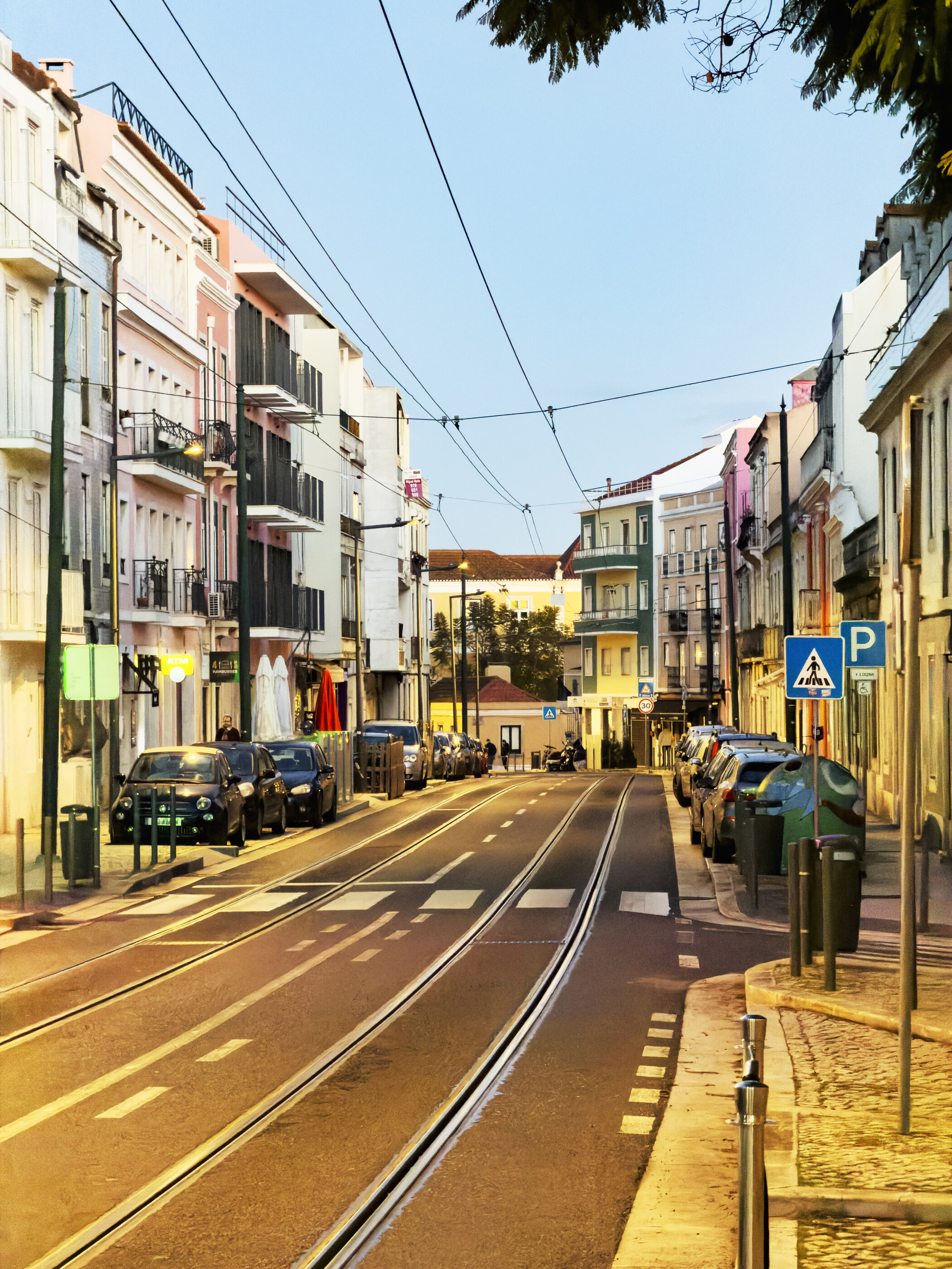 A wide urban street scene with cars, tram tracks, and tall buildings on both sides under bright daylight.